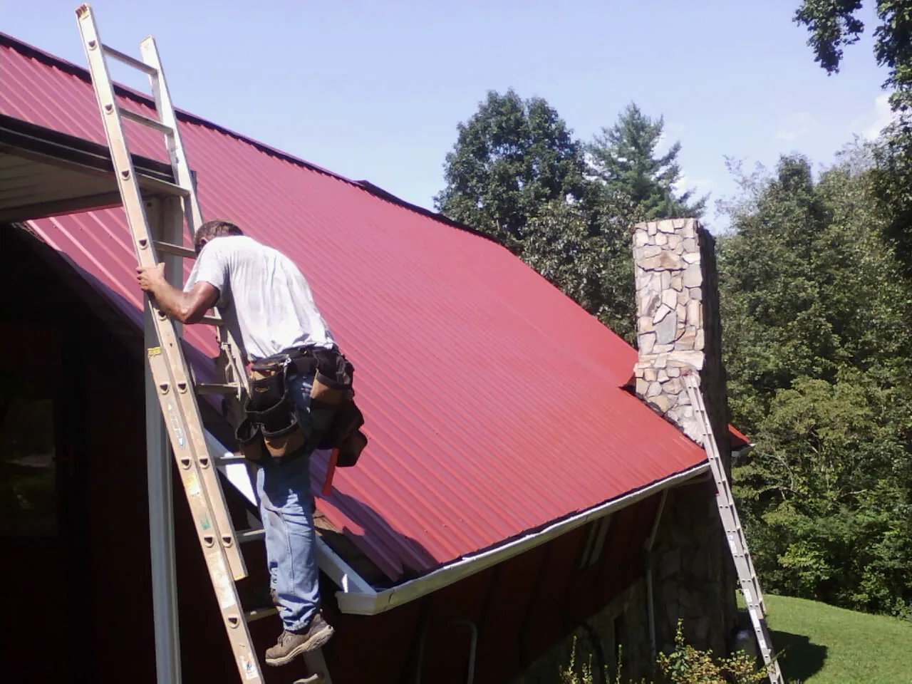 Roofer on a ladder beside a red metal roof and stone chimney — Soffit & Fascia Repair by Roofing Brothers in Gallipolis